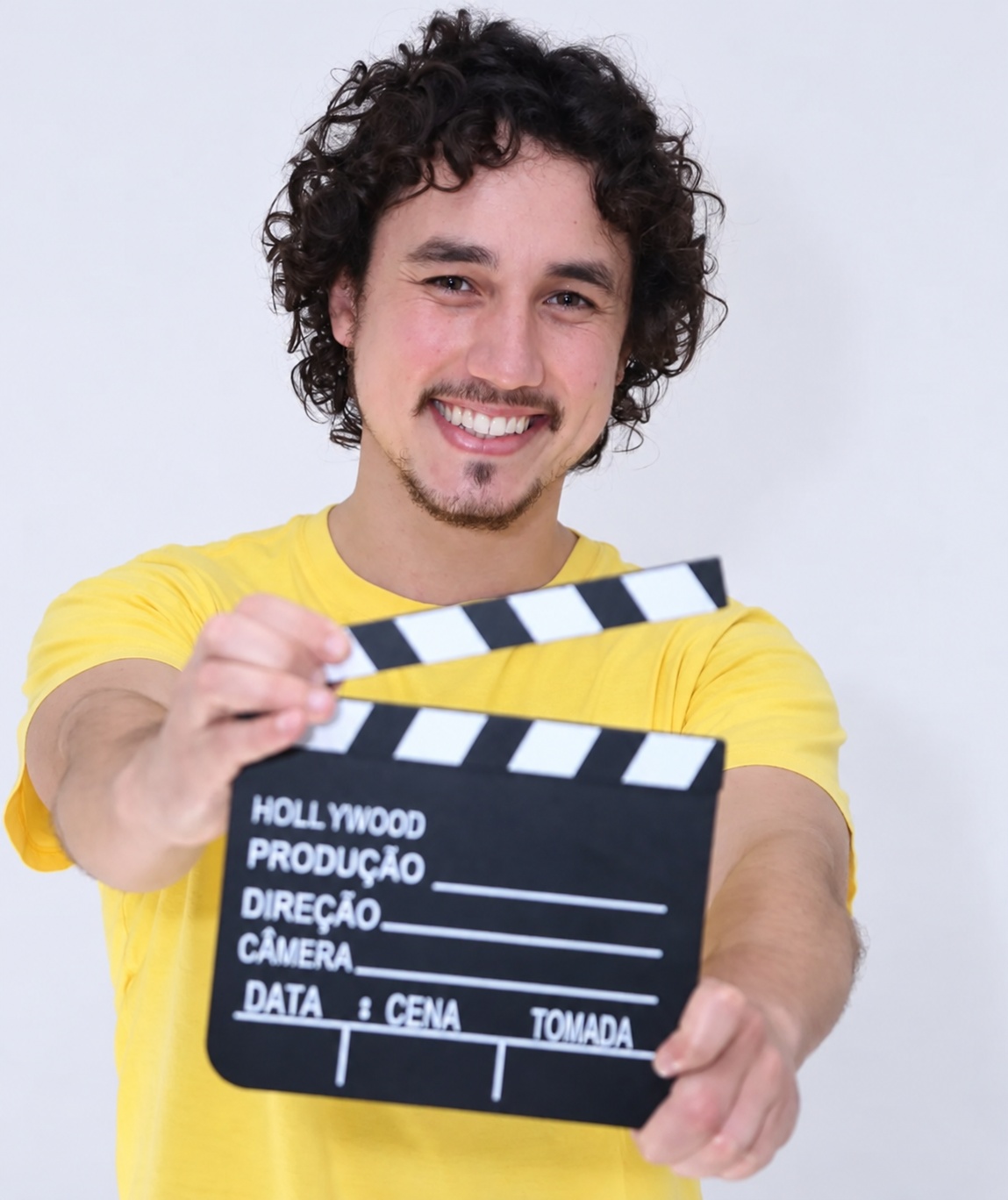 João Paulo sorrindo, de camiseta amarela, segurando uma claquete de cinema